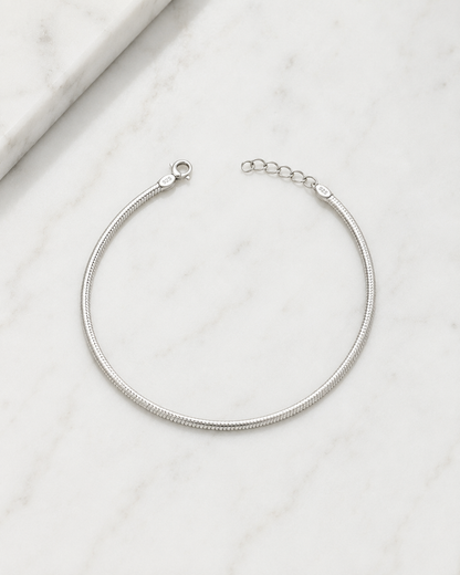A silver snake chain bracelet with a lobster clasp and extension chain, displayed on a white marble surface.